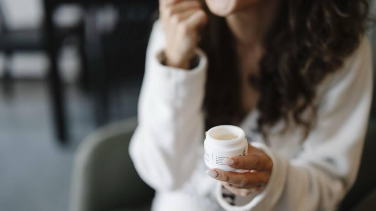 Femme tenant un petit pot de crème blanche ouvert, assise à une table